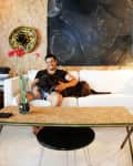 Man sitting on a white sofa with a dog, in front of a chalkboard and decorative plate, with flowers on a table.