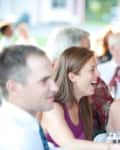 People sitting at a table, smiling and laughing, holding drinks at an outdoor event.