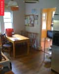 Small kitchen with wooden table, chairs, bulletin board, and stainless steel fridge. Red bag on table, doorway to bright room.