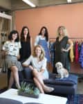 Five women and a dog in a room with a peach wall, clothing rack, and dark sofa with succulents on a table.