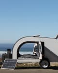 Teardrop camper with open hatch and solar panel on a beach, ocean in background, people walking nearby.
