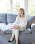 Woman in white sweater and light jeans sitting on a blue sofa, smiling, with black heels and a gold watch.