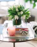 Glass coffee table with pink candle, white tulips in a vase, and decorative bowls in a bright living room.