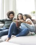 Family reading together on a gray sofa by large windows, with a book in the mother's hands.
