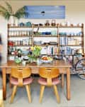 Cozy kitchen with wooden table, colorful chairs, open shelves, and a bicycle. Woman in blue dress walking by.