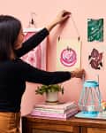 Woman hanging art on a pink wall above a wooden dresser with plants, books, a blue lantern, and a gold lamp.