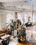 Loft-style studio with exposed brick, large windows, plants, yellow sofa, and art supplies on wooden floors.