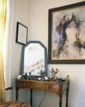 Wooden vanity with makeup, brushes, and a mirror, next to a colorful round stool and abstract portrait on the wall.