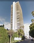 Tall white high-rise building surrounded by palm trees and greenery on a sunny day.