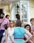 Group of people socializing on a porch with string lights and a yellow house in the background.