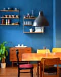 Dining room with blue walls, wooden table, mixed chairs, pendant light, shelves with dishes, and a potted plant.