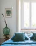 Bedroom with teal bedding, large green vase on a wooden stool, and a framed artwork near a window.