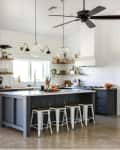 Modern kitchen with stainless steel fridge, island with stools, pendant lights, and open shelves with plants and decor.