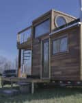 Tiny house on wheels with wood siding, large windows, and a rooftop deck with spiral staircase.