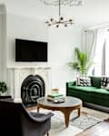 Living room with green velvet sofa, black armchairs, white fireplace, large window, and modern chandelier.