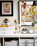 Home office desk with organized storage boxes, framed art, corkboard with notes, and decorative jars.