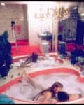 Couple in a heart-shaped bathtub with bubbles, surrounded by mirrored walls and a chandelier above.