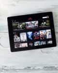 Tablet displaying Netflix interface on a wooden table with a cup of coffee and white earphones.