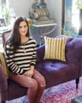 Woman sitting on a purple sofa with striped pillows, wearing a striped sweater and ripped jeans, on a colorful rug.