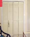 Cream-colored louvered closet doors with a black cord hanging, surrounded by light walls and a cluttered floor.