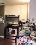 Cluttered kitchen with stainless steel fridge, white cabinets, and various items on countertops.
