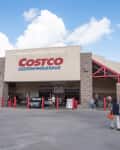 Costco Wholesale store entrance with people walking, red accents, and a clear sky.