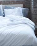Unmade bed with white sheets and pillows against a brick wall, small plant on bedside table.