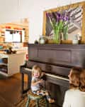 Child smiling while sitting at a dark wooden piano, with a vase of purple flowers on top.