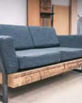 Gray sofa with metal frame and wooden base in a workshop setting, surrounded by tools and boxes.