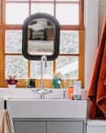 Bathroom sink with a mirror, orange towel, potted plants, and toiletries on the windowsill.