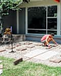 Person in red tank top arranging bricks on gravel in front of large windows.