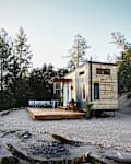 Tiny house with wooden siding on a gravel lot, surrounded by trees, featuring a small deck with outdoor seating.