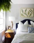 Bedroom with a navy headboard, white bedding, wooden nightstand, lamp, hanging fern, and bird artwork on the wall.