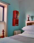 Bedroom with light blue walls, red wall shelves, white bedding, and a red lamp on a white headboard.