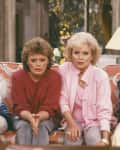 Four women sitting on a floral sofa, focused on a TV, with a lamp and flowers in the background.