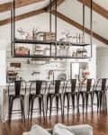 Rustic kitchen with exposed beams, metal bar stools, open shelving, and a large island under hanging shelves.