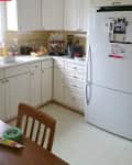 Corner kitchen with white cabinets, fridge, espresso machine, and fruit bowl on counter. Brown chair and table with tape and caulk.