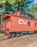 Red caboose with "CV" lettering on train tracks, surrounded by trees and blue sky.
