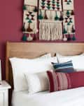 Bedroom with a wooden headboard, red accent pillows, geometric wall hanging, and white nightstands with lamps.