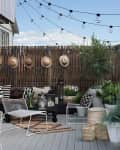 Outdoor patio with string lights, wicker fence, potted plants, white chairs, and hats hanging on the wall.