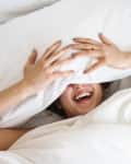 Person laughing under white bedding, covering face with pillow, showing hands with painted nails.