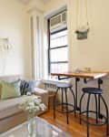 Cozy living room with beige sofa, green and blue pillows, wall art, and a small wooden table with two stools by the window.