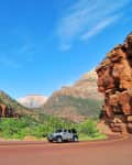 Jeep driving on a winding road through a canyon with red rock cliffs and green vegetation under a clear blue sky.