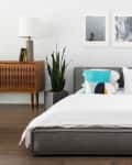 Modern bedroom with a gray bed, colorful pillows, wooden sideboard, and yellow lamp, featuring framed art above the bed.
