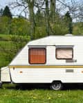 White caravan with brown curtains parked on a grassy field surrounded by trees.