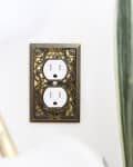 Decorative brass outlet cover with intricate floral and scroll design on a white wall.