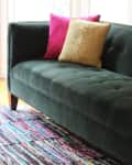Green velvet sofa with pink and gold cushions on a colorful striped rug.
