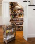 Under-stair pantry with open doors, shelves of jars, cans, and baskets, and a wooden cart with jars and boxes on a wooden floor.