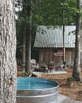 Rustic cabin with metal roof, surrounded by trees, Adirondack chairs, fire pit, and a metal tub in the foreground.