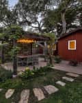 Garden patio with string lights, wooden pergola, and outdoor dining set next to a red cabin surrounded by lush greenery.